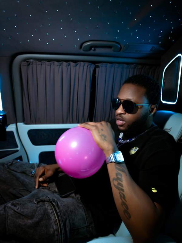 A Man In Sunglasses Sitting In A Car With A Star Patterned Ceiling Light, Holding A Pink Balloon.